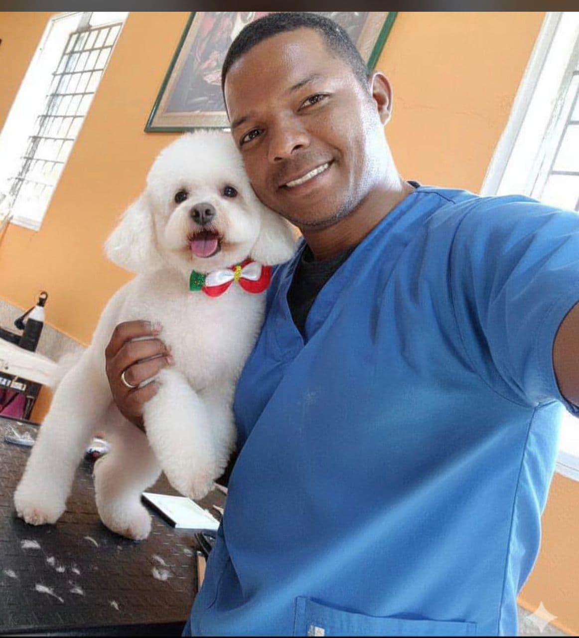 Hombre sonriente con uniforme azul abraza a un perro blanco esponjoso con un lazo festivo.